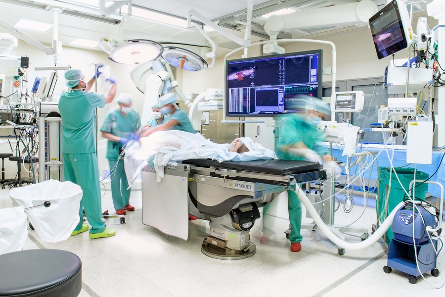 Foto von einem OP-Saal mit Patient auf OP-Tisch, medzinischen Geräten und 5 Personen in grüner OP-Kleidung  Foto von einem OP-Saal mit Patient auf OP-Tisch, medzinischen Geräten und 5 Personen in grüner OP-Kleidung