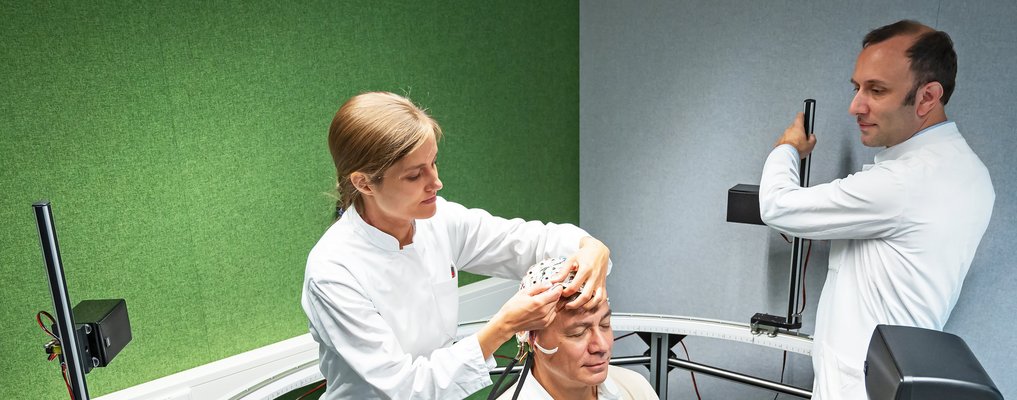 Eine Frau setzt einem Patienten eine Haube mit Leitungen auf en Kopf. ein Arzt stellt die Geräte für eine Messung ein