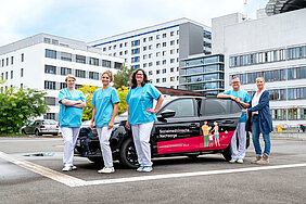 Ein schwarzer Kleinwagen mit Beschriftung „Sozialmedizinische Nachsorge“ steht auf einem Platz, im Hintergrund ist das Universitätsklinikum Halle (Saale) zu sehen. Neben dem Auto stehen drei Frauen und ein Mann in medizinischer Arbeitskleidung sowie eine Frau in Zivil.