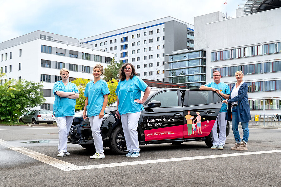 Ein schwarzer Kleinwagen mit Beschriftung „Sozialmedizinische Nachsorge“ steht auf einem Platz, im Hintergrund ist das Universitätsklinikum Halle (Saale) zu sehen. Neben dem Auto stehen drei Frauen und ein Mann in medizinischer Arbeitskleidung sowie eine Frau in Zivil.  Ein schwarzer Kleinwagen mit Beschriftung „Sozialmedizinische Nachsorge“ steht auf einem Platz, im Hintergrund ist das Universitätsklinikum Halle (Saale) zu sehen. Neben dem Auto stehen drei Frauen und ein Mann in medizinischer Arbeitskleidung sowie eine Frau in Zivil.