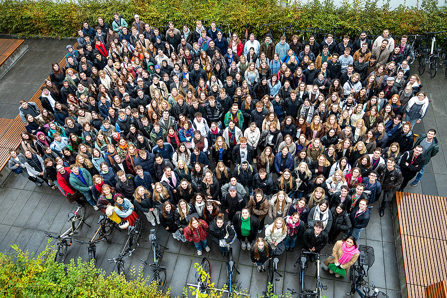 Das Gruppenbild aus der Vogelperspektive zeigt mehrere hundert Personen, die nach oben schauen. An dem nassen Boden und den dicken Jacken kann man erkennen, dass es zuvor geregnet hat. Im Hintergrund sind Büsche und Fahrräder zu sehen.  Das Gruppenbild aus der Vogelperspektive zeigt mehrere hundert Personen, die nach oben schauen. An dem nassen Boden und den dicken Jacken kann man erkennen, dass es zuvor geregnet hat. Im Hintergrund sind Büsche und Fahrräder zu sehen.