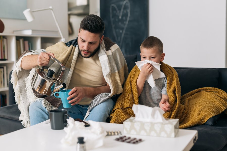 Ein Mann und ein Junge sitzen auf einem Sofa. Beide haben eine warme Decke um die Schultern. Der Mann gießt heißes Wasser aus einer Teekanne in eine Tasse. Der Junge putzt sich die Nase. Auf dem Tisch vor den beiden liegen verschiedene Tablettenschachteln und Taschentücher. Im Hintergrund sind ein Bücherregal, eine Lampe und ein Spiegel zu erkennen.  Ein Mann und ein Junge sitzen auf einem Sofa. Beide haben eine warme Decke um die Schultern. Der Mann gießt heißes Wasser aus einer Teekanne in eine Tasse. Der Junge putzt sich die Nase. Auf dem Tisch vor den beiden liegen verschiedene Tablettenschachteln und Taschentücher. Im Hintergrund sind ein Bücherregal, eine Lampe und ein Spiegel zu erkennen.