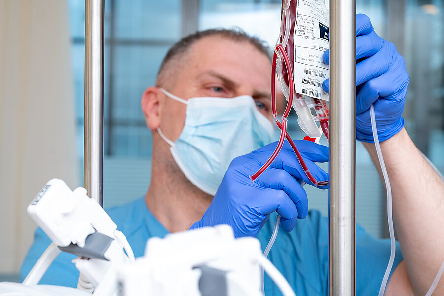 Eine Person in medizinischer Arbeitskleidung hält einen Blutbeutel für eine Stammzelltransplantation in den Händen. Die Person trägt eine Maske und Handschuhe.  Eine Person in medizinischer Arbeitskleidung hält einen Blutbeutel für eine Stammzelltransplantation in den Händen. Die Person trägt eine Maske und Handschuhe.