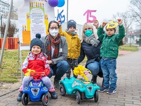 Die Pflegedirektorin Christiane Becker und die stellv. Kita-Leiterin Daniela Werner hocken neben drei Kindern. Ein Kind sitzt auf einem blaue Bobbycar, ein türkisfarbenes steht daneben. Daniela Werner und die zwei anderen Kinder halten die Zahl 15 aus Papier in die Luft.