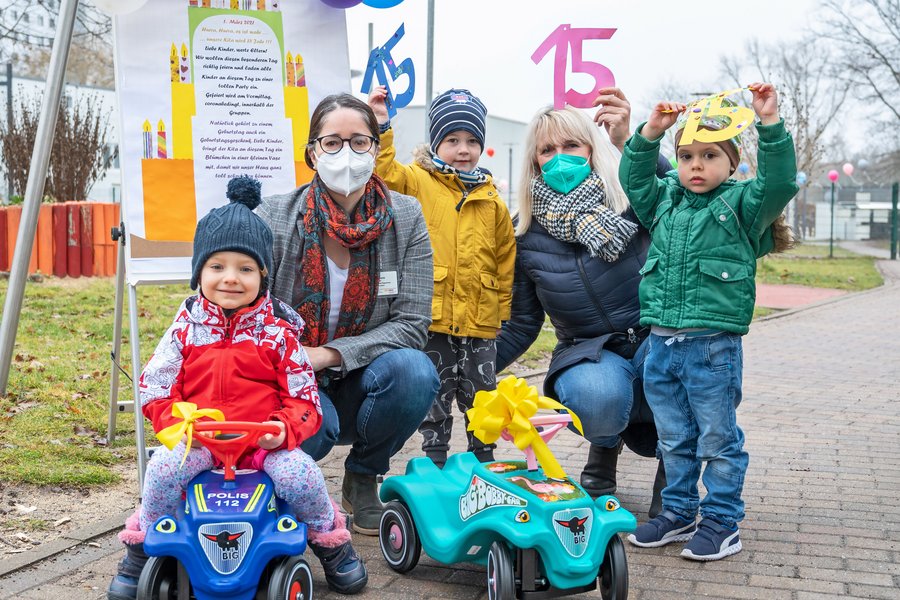 Die Pflegedirektorin Christiane Becker und die stellv. Kita-Leiterin Daniela Werner hocken neben drei Kindern. Ein Kind sitzt auf einem blaue Bobbycar, ein türkisfarbenes steht daneben. Daniela Werner und die zwei anderen Kinder halten die Zahl 15 aus Papier in die Luft.  Die Pflegedirektorin Christiane Becker und die stellv. Kita-Leiterin Daniela Werner hocken neben drei Kindern. Ein Kind sitzt auf einem blaue Bobbycar, ein türkisfarbenes steht daneben. Daniela Werner und die zwei anderen Kinder halten die Zahl 15 aus Papier in die Luft.