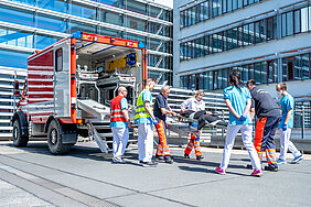 Einsatzkräfte der Feuerwehr und Mitarbeitende der Universitätsmedizin Halle versorgen eine Person auf einer Trage und schieben sie in ein Rettungsfahrzeug der Feuerwehr. 
