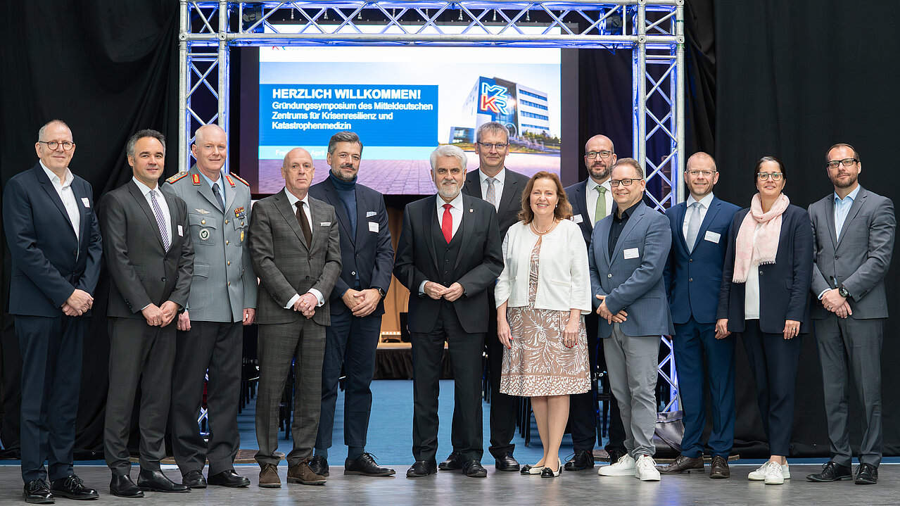 Gruppe von Personen steht auf Bühne vor Bildschirm beim Gründungssymposium des Zentrums für Krisenresilienz und Katastrophenmedizin.