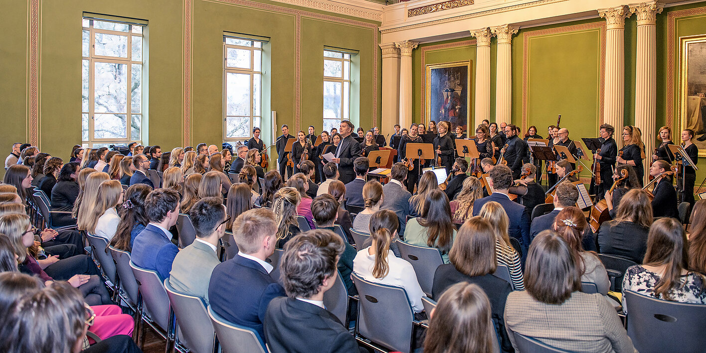 Blick in die Aula des Löwengebäudes der Universität Halle. Der Raum ist gefüllt mit sitzenden Menschen. Am rechten Bildrand, im vorderen Bereich des Raumes, sind zahlreiche Musiker:innen mit ihren Instrumenten zu erkennen.