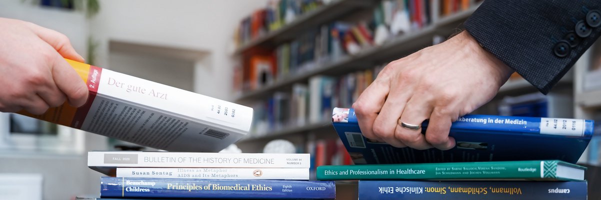 zwei Menschen greifen nach Büchern auf dem Tisch, im Hintergrund sind Bücherregale