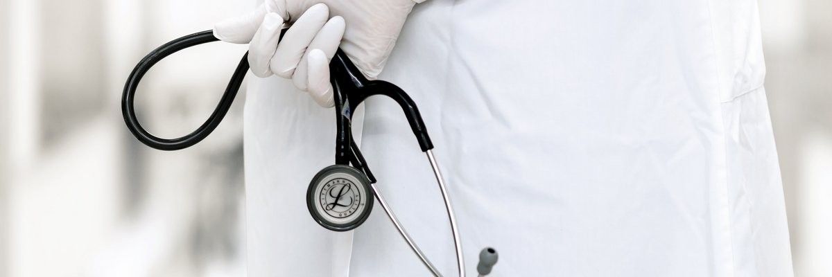 Arzt mit Handschuhen verschränkt die Hände hinter dem Rücken, in denen er ein Stethoskop hält.