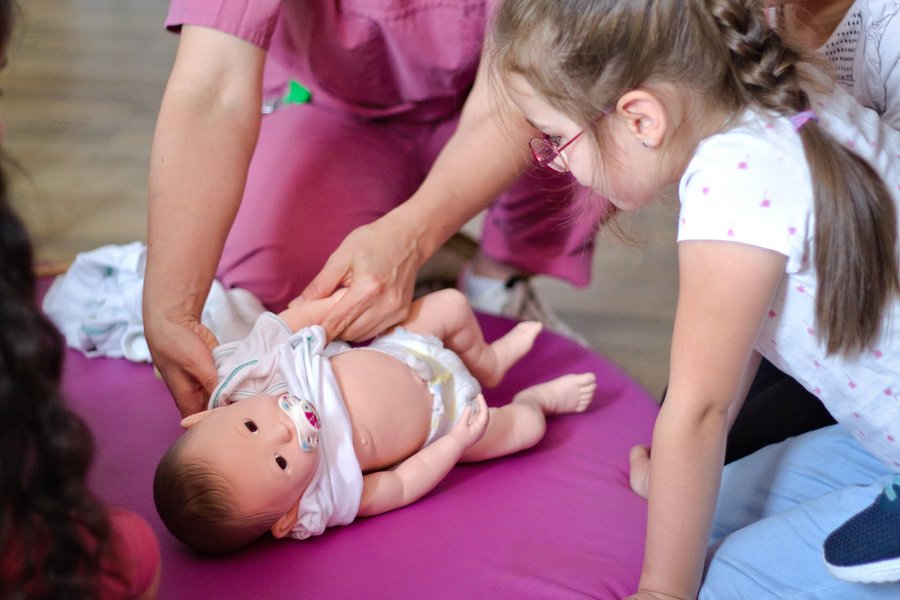 Ein Mädchen schaut aufmerksam auf eine Babypuppe, die auf einer Unterlage auf dem Boden liegt. Eine Person in beerenfarbener medizinischer Arbeitskleidung zieht der Puppe ein Jäckchen an.  Ein Mädchen schaut aufmerksam auf eine Babypuppe, die auf einer Unterlage auf dem Boden liegt. Eine Person in beerenfarbener medizinischer Arbeitskleidung zieht der Puppe ein Jäckchen an.