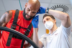 Ein Sportler in rotem Trikot wird auf einem Laufbandstehen von einem Sportwissenschaftler Blut am Ohr (Laktattest) abgenommen. Beide Männer sind ab der oberen Körperhälfte zu sehen.