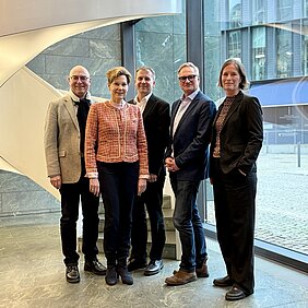 Fünf Personen stehen nebeneinander in einem Raum. Im Hintergrund ist eine beleuchtete, spiralförmige Treppe zu erkennen. Der Gebäudeteil auf der rechten Seite besteht vollständig aus Glas.