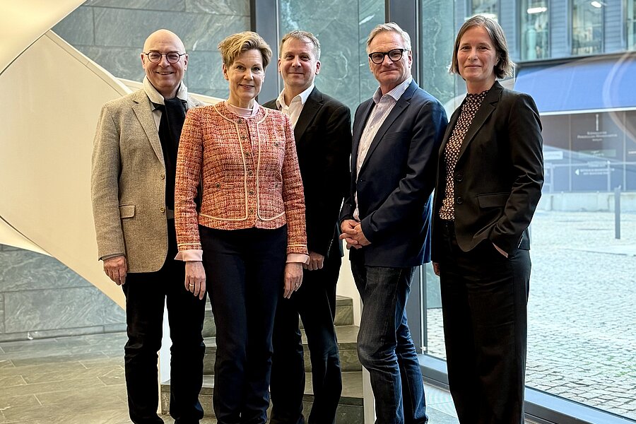 Fünf Personen stehen nebeneinander in einem Raum. Im Hintergrund ist eine beleuchtete, spiralförmige Treppe zu erkennen. Der Gebäudeteil auf der rechten Seite besteht vollständig aus Glas.  Fünf Personen stehen nebeneinander in einem Raum. Im Hintergrund ist eine beleuchtete, spiralförmige Treppe zu erkennen. Der Gebäudeteil auf der rechten Seite besteht vollständig aus Glas.