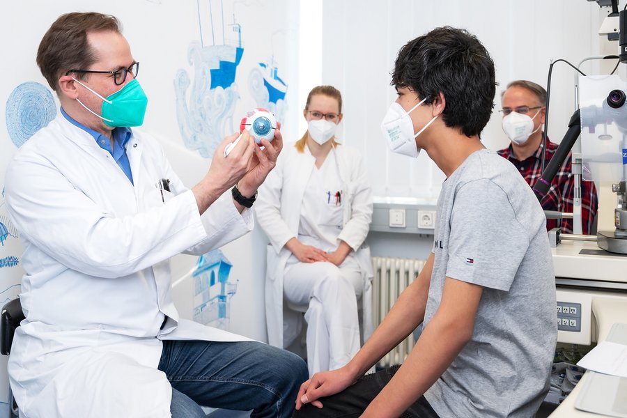 Links im Bild ist Prof. Dr. Arne Viestenz zu sehen. Er hält ein Augenmodell in der Hand und zeigt mit einem Stift darauf. Ihm gegenüber sitzt Komail. Im Hintergrund sitzen Assistenzärztin Christiane Kesper und Maik Dawedeit und schauen zu.  Links im Bild ist Prof. Dr. Arne Viestenz zu sehen. Er hält ein Augenmodell in der Hand und zeigt mit einem Stift darauf. Ihm gegenüber sitzt Komail. Im Hintergrund sitzen Assistenzärztin Christiane Kesper und Maik Dawedeit und schauen zu.