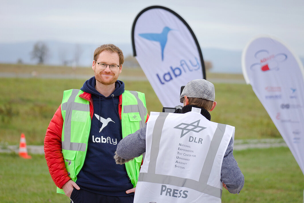 Ein Mann mit Bart und Brille wird von einem Kameramann interviewt. In Hintergrund stehen Beachflags mit Logos auf einer Wiese.