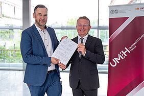 Zwei Männer in Änzügen stehen vor einem Roll-up mit dem Logo der UMH. Sie halten gemeinsam ein Schriftstück in die Kamera.