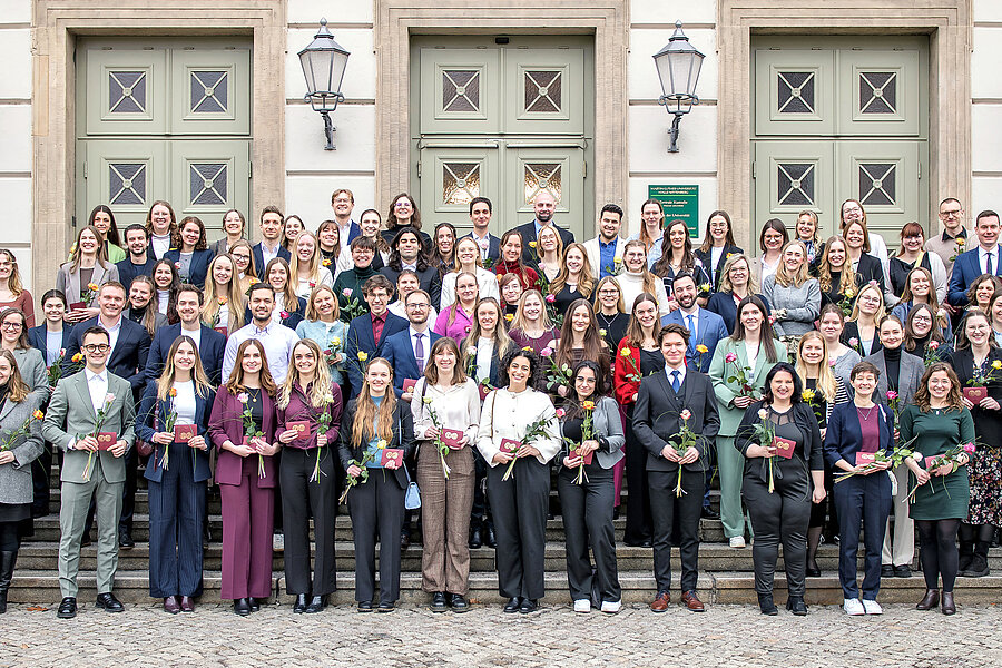 94 Personen stehen auf der Treppe vor dem Löwengebäude der Universität Halle-Wittenberg und lächeln in die Kamera. Sie halten jeweils ein kleines, dunkelrotes Couvert mit einem goldenen Siegel der Universität sowie eine orange-rote Rose in den Händen.  94 Personen stehen auf der Treppe vor dem Löwengebäude der Universität Halle-Wittenberg und lächeln in die Kamera. Sie halten jeweils ein kleines, dunkelrotes Couvert mit einem goldenen Siegel der Universität sowie eine orange-rote Rose in den Händen.