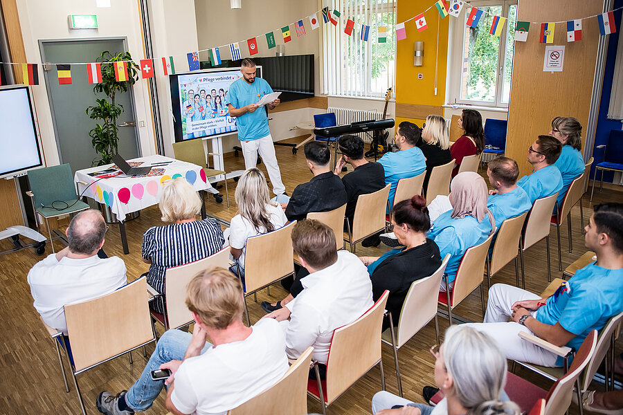 Teilnehmende eines Aktionstages sitzen in einem Raum. Vorne steht eine Person und erzählt etwas. Der Raum ist mit Girlanden aus Fahnen verschiedener Länder geschmückt.   Teilnehmende eines Aktionstages sitzen in einem Raum. Vorne steht eine Person und erzählt etwas. Der Raum ist mit Girlanden aus Fahnen verschiedener Länder geschmückt. 