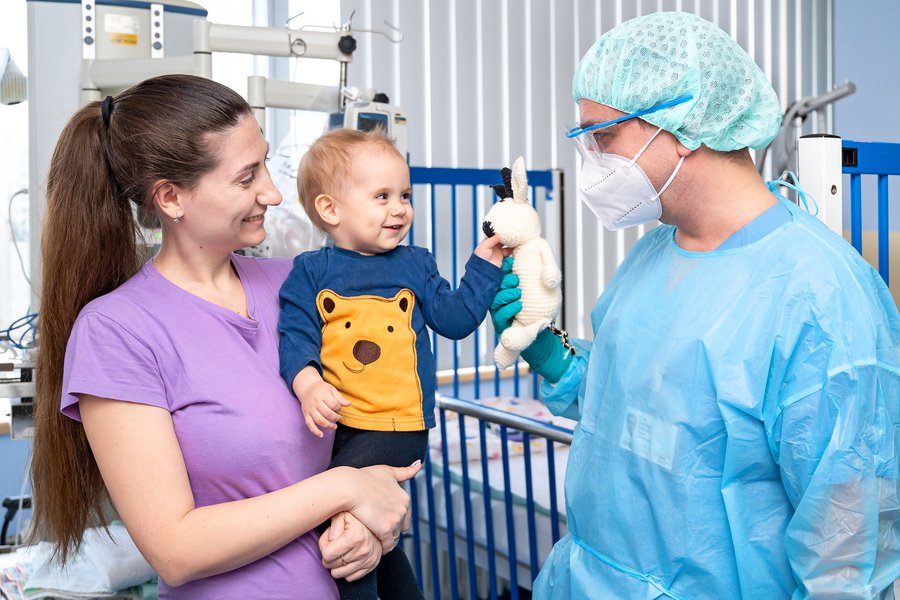 Eine junge Frau hat ein kleines Kind auf dem Arm. Ein Arzt in medizinscher Schutzkleidung steht rechts von ihnen und reicht dem Kind einen teddybär.  Eine junge Frau hat ein kleines Kind auf dem Arm. Ein Arzt in medizinscher Schutzkleidung steht rechts von ihnen und reicht dem Kind einen teddybär.