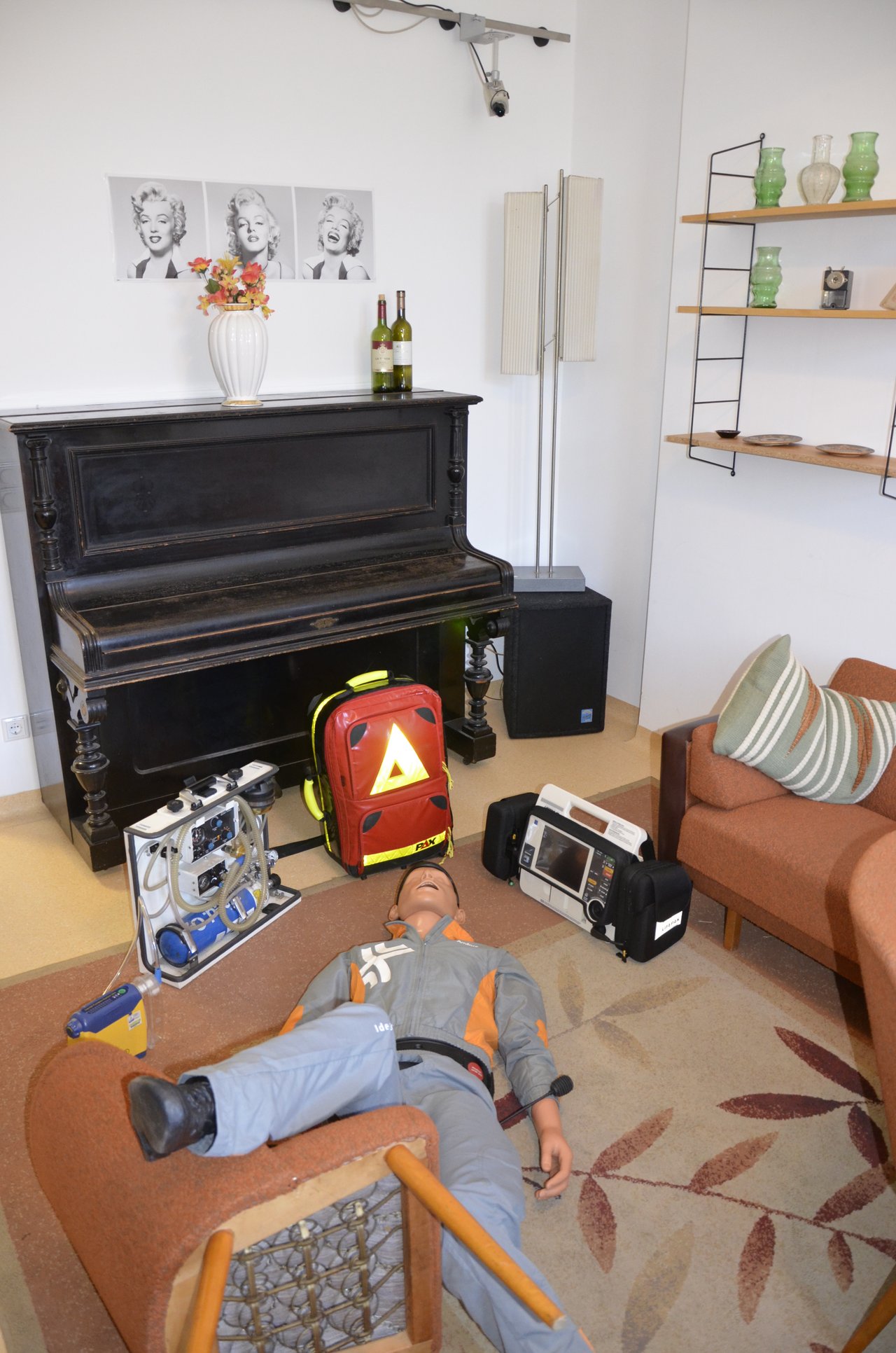 Eine Simulations-Puppe liegt auf dem Boden eines Wohnzimmers; ein Bein liegt über einem umgefallenen Sessel. Daneben stehen Notarzt-Koffer und -Utensilien.