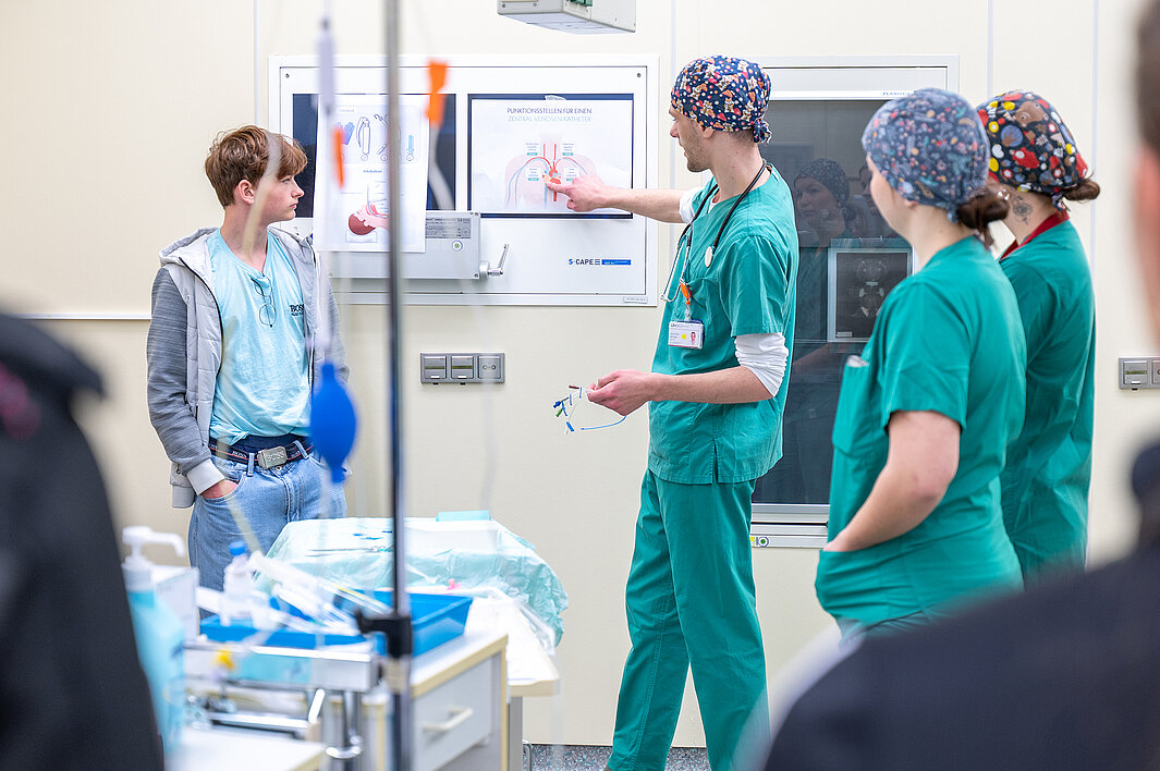 Rechts im Bild stehen drei Personen in OP-Kleidung. Eine der Personen erklärt einem Schüler etwas anhand eines Schaubildes.