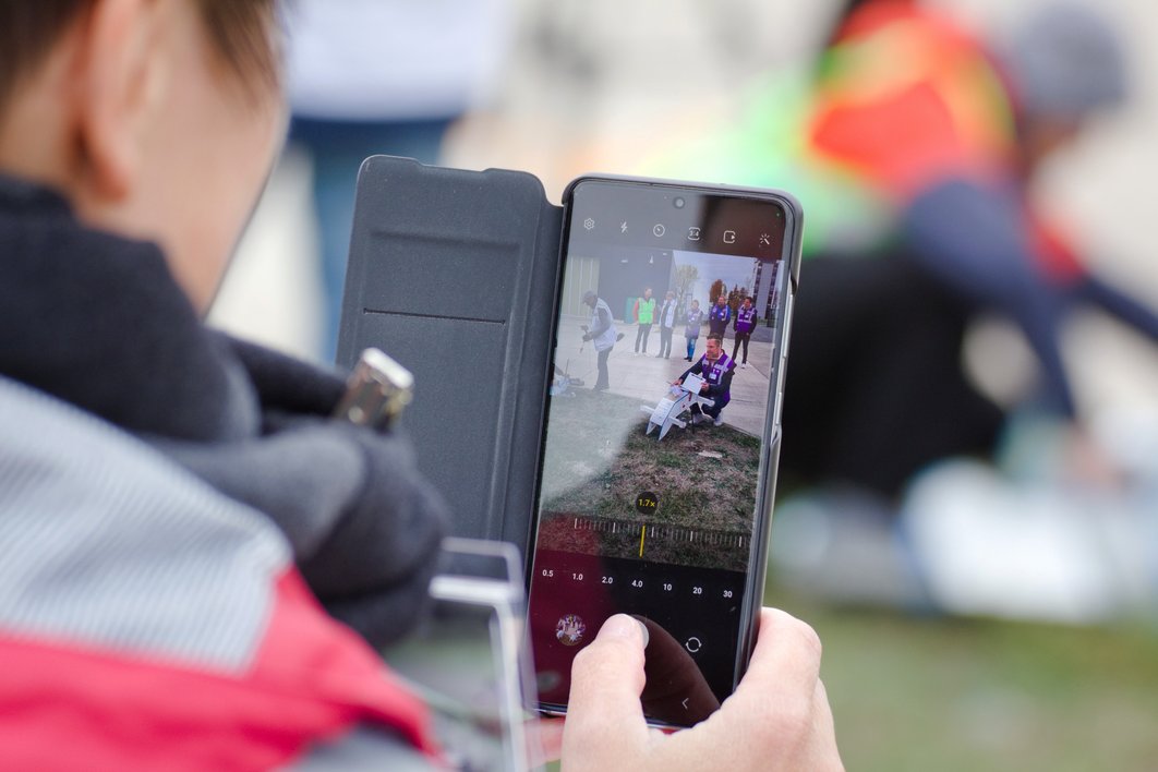 Eine Frau macht ein Foto mit dem Handy. Das Bild zeigt das Display während der Aufnahme. Darauf sind Menschen und die Flugdrohne zu erkennen.
