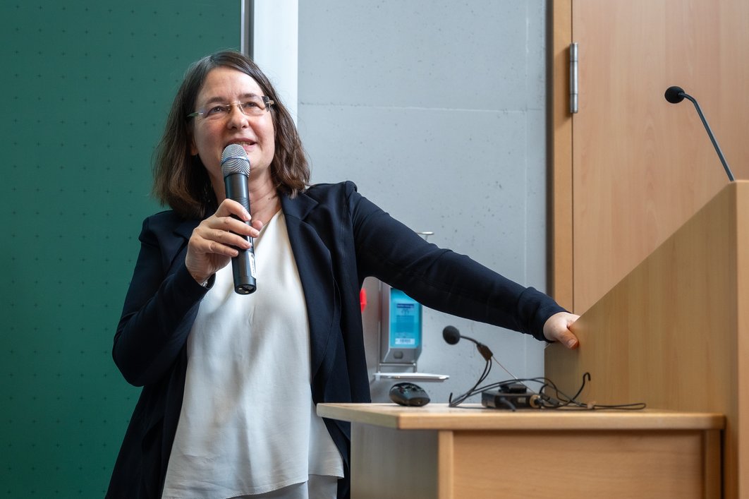Eine Person in weißer Bluse und schwarzem Blazer steht mit Mikro in der Hand locker an einem Redepult.