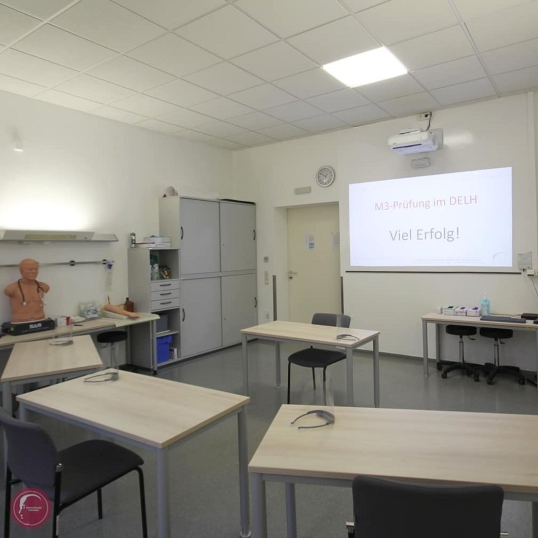 Tische in einem Seminarraum; auf jedem Tisch liegt eine 3D-Brille; an die Wand ist projiziert: "M3-Prüfung im DELH - Viel Erfolg!"