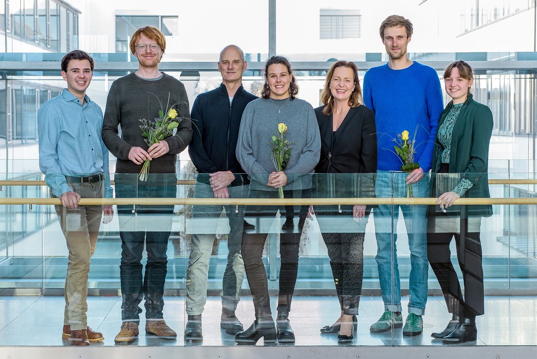 Sieben Personen stehen in einem lichtdurchfluteten Korridor und lächeln in die Kamera. Drei halten einen Blumenstrauß.