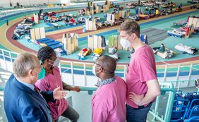 Drei Männer und eine Frau in medizinscher Arbeitskleidung (brombeerfarben) sind im Gespräch zu sehen. Im Hintergrund ist eine Sporthalle mit Feldbetten zu sehen.