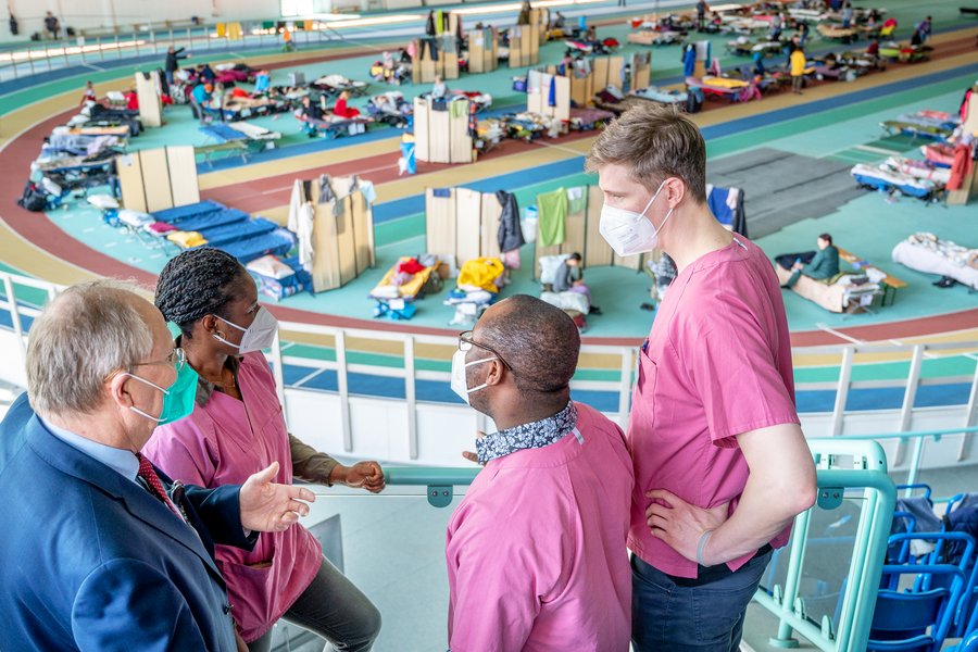 Drei Männer und eine Frau in medizinscher Arbeitskleidung (brombeerfarben) sind im Gespräch zu sehen. Im Hintergrund ist eine Sporthalle mit Feldbetten zu sehen.  Drei Männer und eine Frau in medizinscher Arbeitskleidung (brombeerfarben) sind im Gespräch zu sehen. Im Hintergrund ist eine Sporthalle mit Feldbetten zu sehen.