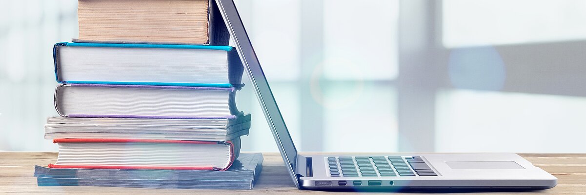 Stapel Bücher auf einem Tisch, ein Laptop lehnt an den Büchern