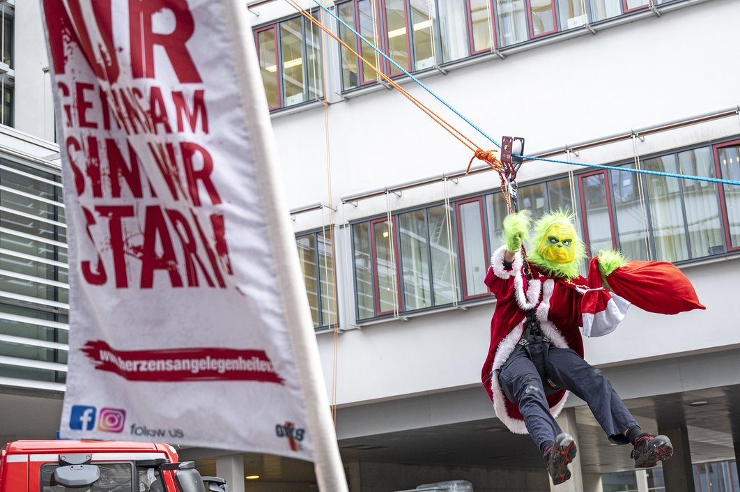 Ein als Grinch verkleideter Mitarbeiter der Feuerwehr seilt sich vom Universitätsklinikum Halle (Saale) ab. Links steht eine Fahne mit der Aufschrift "Nur gemeinsam sind wir stark".