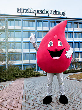 Das Maskottchen der Blutspende des Universitätsklinikums Halle (Saale), ein großer Blutstropfen mit Gesicht, steht vor dem Verlagsgebäude der MZ in Halle (Saale). Es handelt sich um ein mehrstöckiges Bürogebäude. Auf dem Dach steht der Schriftzug „Mitteldeutsche Zeitung“.