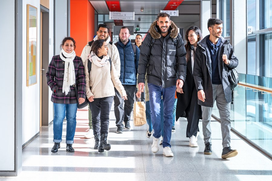 Eine Gruppe junger Männer und Frauen läuft gut gelaunt einen Gang mit Glasfront entlang.  Eine Gruppe junger Männer und Frauen läuft gut gelaunt einen Gang mit Glasfront entlang.