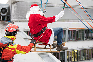 Ein Weihnachtsmann sitzt auf einem Holzschlitten und hängt an einem Sicherungsseil. Er steht kurz vor der Abfahrt an einem Seil herunter von einem Gebäudedach, ein Mitarbeiter der Höhenrettung der Feuerwehr hält ihn fest.