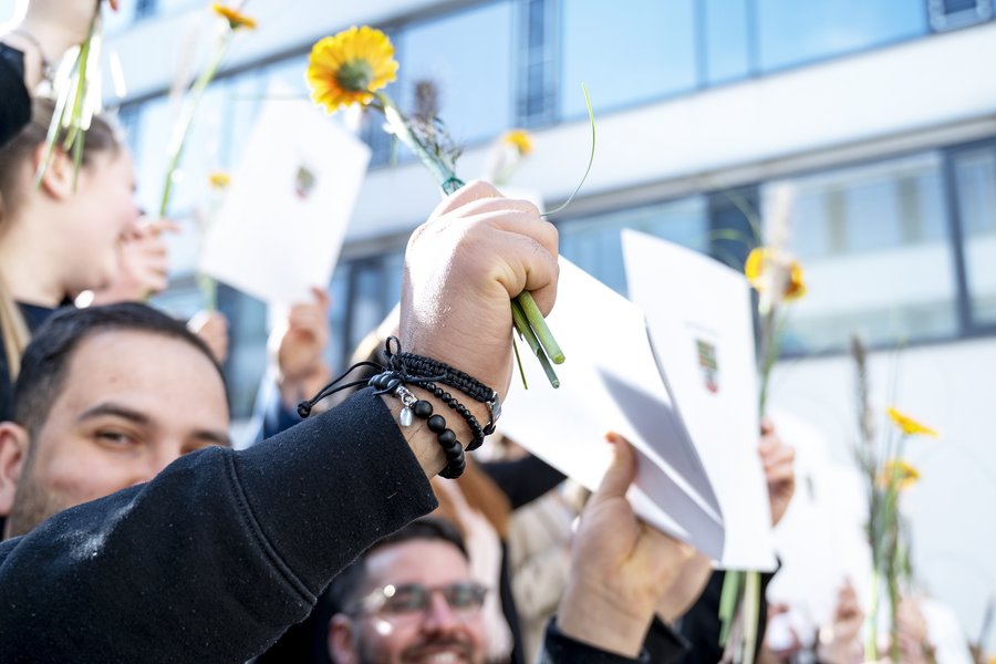 Seitlicher Blick auf Absolvent:innen, die Zeugnisse und Blumen in die Luft strecken.  Seitlicher Blick auf Absolvent:innen, die Zeugnisse und Blumen in die Luft strecken.