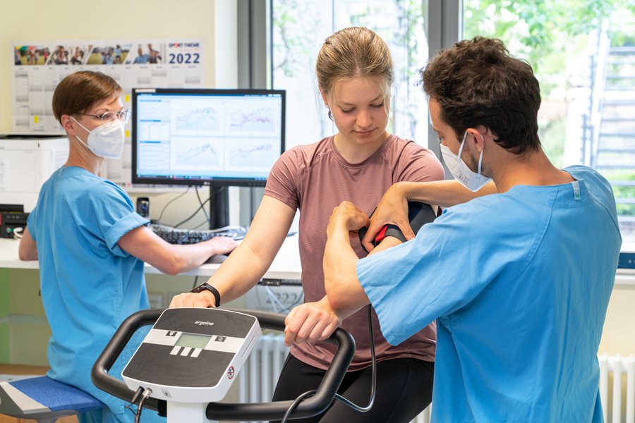Drei Personen befinden sich in einem Trainingsraum. Eine junge Frau sitzt auf einem Heimtrainer und bekommt eine Blutdruckmanschette angelegt. Die dritte Person steht vor einem Monitor, auf dem unscharf mehrere Graphen zu erkennen sind, und blickt nach hinten zu den anderen beiden Personen.  Drei Personen befinden sich in einem Trainingsraum. Eine junge Frau sitzt auf einem Heimtrainer und bekommt eine Blutdruckmanschette angelegt. Die dritte Person steht vor einem Monitor, auf dem unscharf mehrere Graphen zu erkennen sind, und blickt nach hinten zu den anderen beiden Personen.