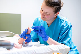 Eine Hebamme hält das Bein eines Babys in der Hand und bemalt den Fuß mit einem Tupfer und goldener Farbe.