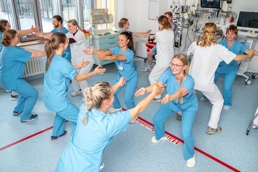 Eine Gruppe von Mitarbeitenden in einem Krankenhaus macht gemeinsam Sportübungen. Die Personen stehen sich gegenüber, haben die Knie gebeugt und die Arme ausgestreckt. In dem Raum sieht man am Rand allerlei medizinische Geräte.  Eine Gruppe von Mitarbeitenden in einem Krankenhaus macht gemeinsam Sportübungen. Die Personen stehen sich gegenüber, haben die Knie gebeugt und die Arme ausgestreckt. In dem Raum sieht man am Rand allerlei medizinische Geräte.