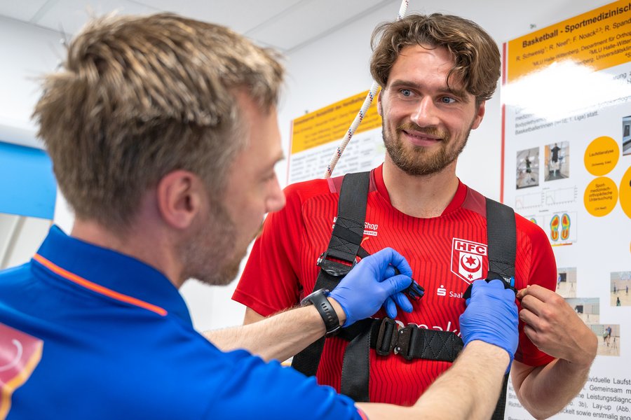 Links ein Mann in blauem Shirt. Rechts Fußballer Jonas Nietfeld in einem roten HFC-Trikot. Er trägt einen Gurt um die Brust, den der andere Mann gerade befestigt.  Links ein Mann in blauem Shirt. Rechts Fußballer Jonas Nietfeld in einem roten HFC-Trikot. Er trägt einen Gurt um die Brust, den der andere Mann gerade befestigt.