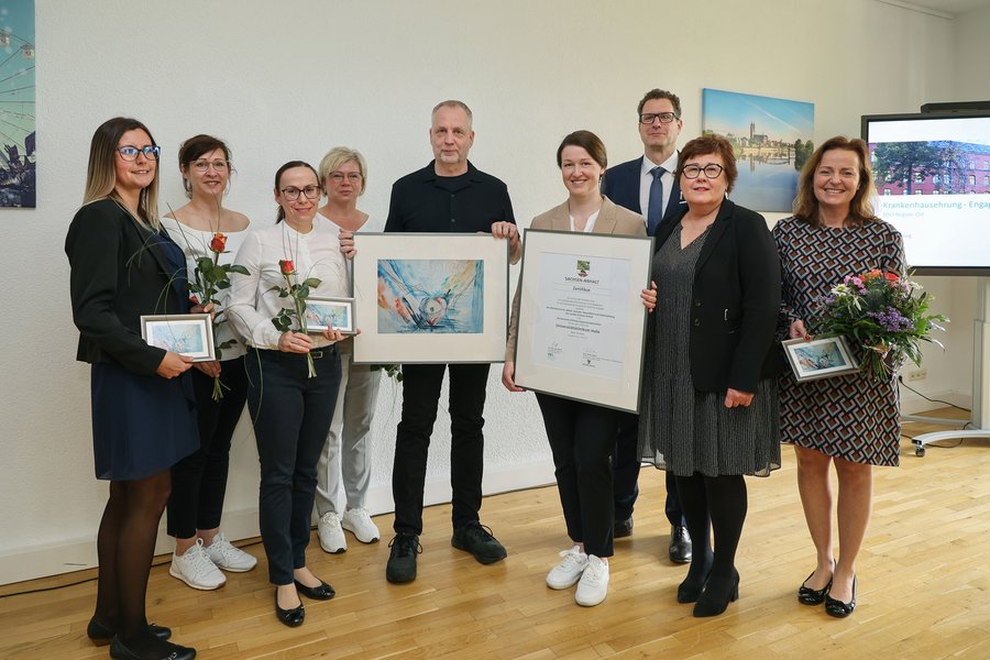 Neun Männer und Frauen stehen in einer Reihe. Sie halten Bilder, Zertifikate und Blumen in den Händen und blicken in die Kamera.  Neun Männer und Frauen stehen in einer Reihe. Sie halten Bilder, Zertifikate und Blumen in den Händen und blicken in die Kamera.