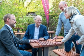 drei Männer und eine Frau sitzen auf Outdoormöbeln und sind im Gespräch. Grüne Bäume im Hintergrund