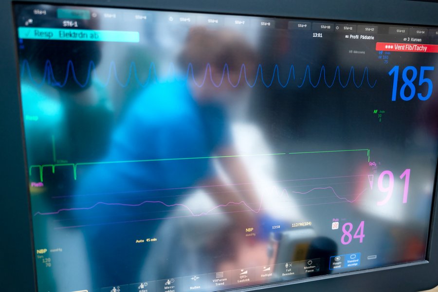 Man sieht die dunkle und matte Oberfläche eines Monitors, der Vitaldaten eines Patienten darstellt. In der Spiegelung des Monitors ist unscharf eine Person zu erkennen, die in blauen medizinischem Arbeitsschutz gekleidet an einem Patienten arbeitet.  Man sieht die dunkle und matte Oberfläche eines Monitors, der Vitaldaten eines Patienten darstellt. In der Spiegelung des Monitors ist unscharf eine Person zu erkennen, die in blauen medizinischem Arbeitsschutz gekleidet an einem Patienten arbeitet.