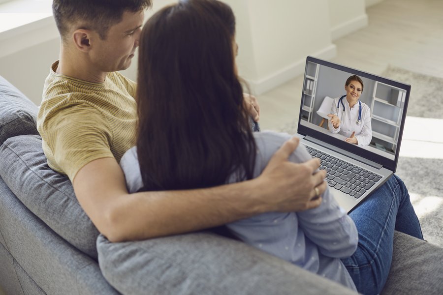 Ein junger Mann sitzt auf einer grauen Couch und hat seinen Arm um die Schultern einer jugen Frau gelegt, die rechts von ihm sitzt. Sie schauen auf den Bildschirm eines Laptops, der vor ihnen auf einem Tisch steht. Auf dem Bilschirm ist eine Ärztin zu sehen.  Ein junger Mann sitzt auf einer grauen Couch und hat seinen Arm um die Schultern einer jugen Frau gelegt, die rechts von ihm sitzt. Sie schauen auf den Bildschirm eines Laptops, der vor ihnen auf einem Tisch steht. Auf dem Bilschirm ist eine Ärztin zu sehen.