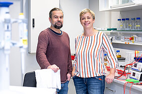 Zwei Personen stehen in einem weißen, hell erleuchteten Labor und lächeln in die Kamera. Links steht ein Mann in einem weinroten Pullover, der seine rechte Hand auf einem Stuhl abstützt. Auf der rechten Seite lehnt eine Frau mit gestreifter Bluse an einem Labortisch. Im Hintergrund und im Vordergrund sind verschiedene Laborgeräte wie Pipetten, Schottflaschen und Gelbäder zu sehen.