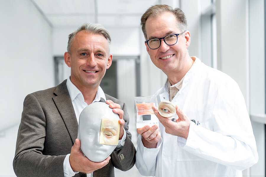 Zwei lächelnde Männer in einem hellen Klinikflur halten anatomische Augenmodelle in die Kamera: ein weißer Kopf mit aufgesetztem Lidmodell, ein transparenter Würfel mit Gewebeprobe und ein rundes Augenmodell mit Lidern.  Zwei lächelnde Männer in einem hellen Klinikflur halten anatomische Augenmodelle in die Kamera: ein weißer Kopf mit aufgesetztem Lidmodell, ein transparenter Würfel mit Gewebeprobe und ein rundes Augenmodell mit Lidern.