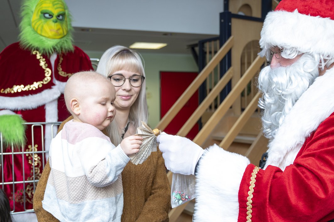 Der Weihnachtsmann gibt einem Kleinkind ein Geschenk. Dahinter befindet sich die Mutter. Im Hintergrund steht der Grinch.