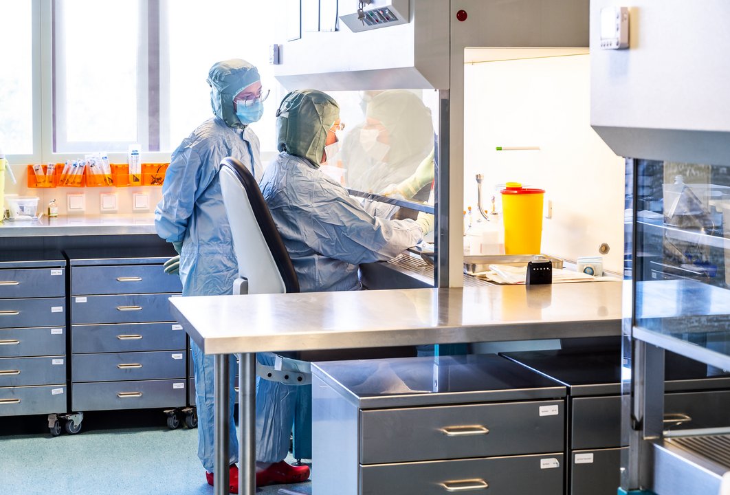 zwei Mitarbeitende im Ganzkörperanzug arbeiten in einem Speziallabor der Universitätsapotheke am Labortisch